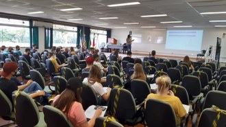 foto de um auditório com várias pessoas sentadas e no fundo um professor dá aula