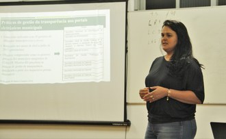 Fotografia em médio plano. Uma mulher em pé, gesticula para o público. Ao seu lado, uma tela branca com projeções de seu trabalho.