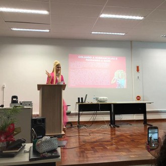 Fotografia em médio plano. Homem está em pé, montado de drag queen (peruca loira, vestido curto rosa pink e maquiado). Ele está em pé falando para pessoas sentadas, dentro de um auditório. Atrás dele, uma tela projetando textos e imagens.