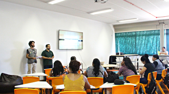 Foto aberta de uma sala, com cadeiras laranjas. Várias mulheres estão sentadas, olhando para frente, onde estão dois homens falando.
