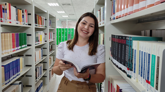 Foto da Luana no corredor da biblioteca, em meio às prateleiras de livros, com um livro aberto. Ela olha para frente e sorri. Luana é uma jovem de pele branca, olhos e cabelos castanhos, usa um piercing no nariz e camiseta branca