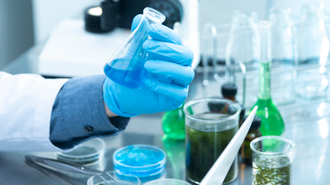 Imagem de laboratório, com um ângulo fechado na mão de uma pessoa. Ela usa luvas e segura um frasco com líquido azul