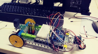 Foto de um protótipo de robô, que tem duas rodas e um chassi, além de fios e pilhas. O protótipo de robô está em cima de uma mesa. Atrás, aparece o teclado de um computador