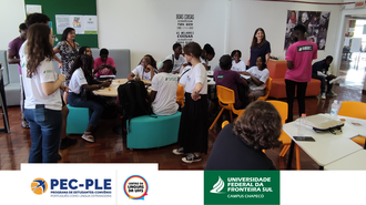 Foto de jovens em uma sala. Eles estão sentados em puffs e cadeiras coloridas ao redor de mesas redondas, e outros estão em pé.