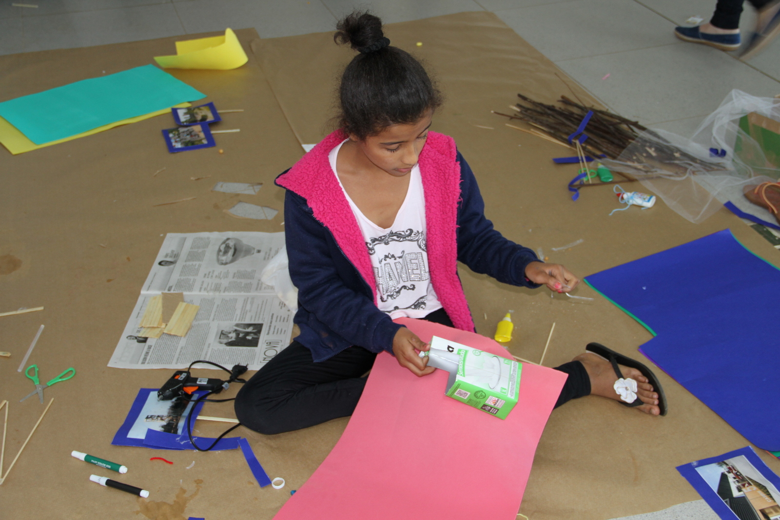 Uma menina, sentada, monta uma maquete sobre uma cartolina com uma caixa de litro de leite