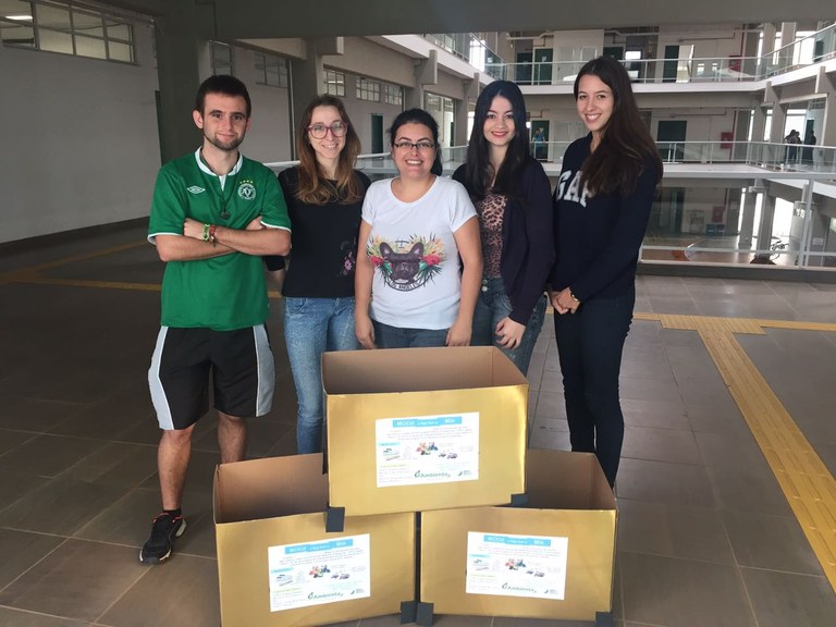Três caixas douradas, duas no chão e a terceira em cima das outras, estão em frente a um homem e quatro mulheres, componentes da Empresa Junior