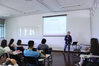 Em pé, na frente do público na sala de aula, uma mulher faz explicações. No fundo, há uma projeção do texto do qual ela está explicando.