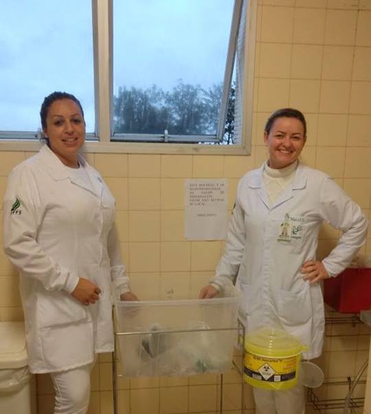 Duas estudantes, vestidas de branco, ao lado da caixa para coleta de material contaminado