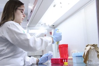 Mulher trabalha de jaleco e luvas no laboratório