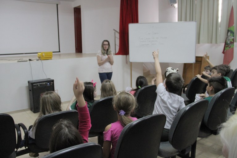 Estudantes em um auditório, de costas, olham para a professora, que explica de frente para a câmera