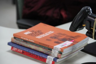 Uma pilha de livros está em cima de uma mesa e, em frente, o leitor de código de barras, cuja luz está apontada para o código fixado em um dos livros