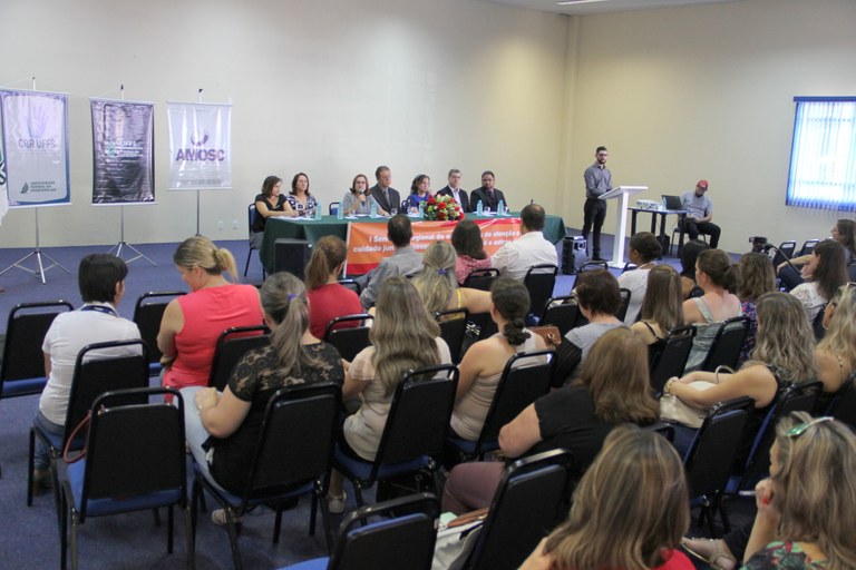 Auditório cheio, com as pessoas sentadas de frente para a mesa de honra, que é formada por sete pessoas