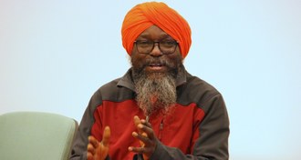 Em plano fechado aparece Dada, um homem de barba branca, com um turbante alaranjado na cabeça. Ele fala e gesticula