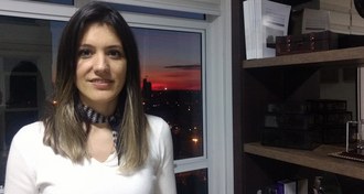 Em plano fechado, foto mostra a médica Camila ao lado de uma estante com livros e, ao fundo, uma janela com o entardecer na cidade