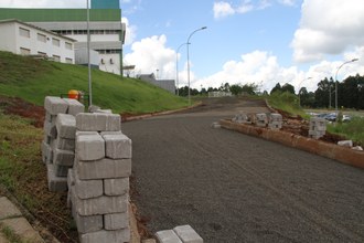 Em primeiro plano, uma pilha de pavers. Ao fundo, o estacionamento apenas com brita. À esquerda, no alto, o Bloco dos Professores.