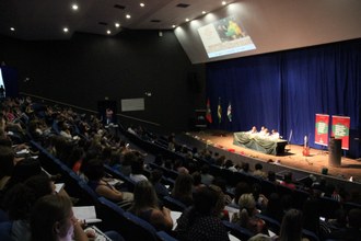 Foto em plano aberto, com o público, de costas para a foto, voltado ao palco, que está mais iluminado, em segundo plano
