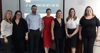 Sete pessoas em pé, em frente a uma tela, com o texto "3º Encontro de NITs de SC" (Foto: Divulgação/UFFS)