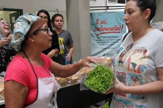 Em plano médio, uma senhora de touca conversa com uma cliente, que segura um pé de alface