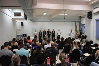 Em plano aberto, o publico aparece em primeiro plano, de costas, olhando para a frente do ambiente, em que estão as autoridades, lado a lado. À frente está um dos médicos falando aos novos residentes