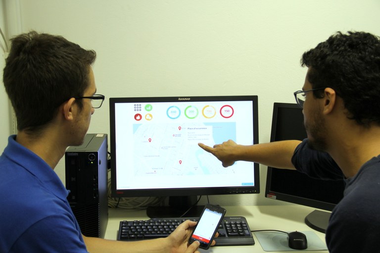 Estudantes discutem sobre o aplicativo em frente a um computador; um deles também segura um smartphone com o aplicativo na tela