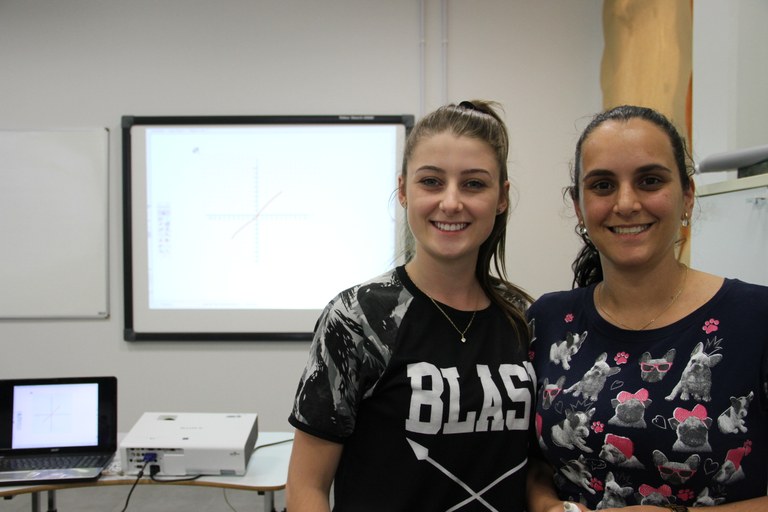 Estudantes, de frente, no canto direito da foto, e, ao fundo, a tela interativa mostra um gráfico