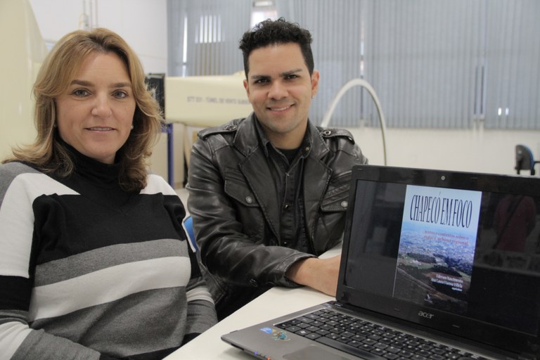 Professora Ana e professor Ederson aparecem atrás de um notebook, no qual aparece a capa do livro