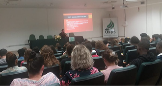Imagem do público de costas, em um auditório. Em cima do palco, sentado atrás de uma mesa, um homem fala ao público.