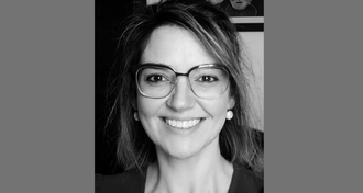 Foto da professora em preto e branco. Ela está em plano fechado, sorrindo e olhando para a câmera