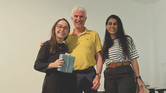 Foto de três pessoas sorridentes olhando para a foto. O homem está no meio, ladeado pelas professoras Sarah, à esquerda, e Debora, à direita