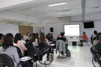 Foto em plano aberto, em um auditório, com pessoas sentadas de costas para a foto e de frente para o palestrante