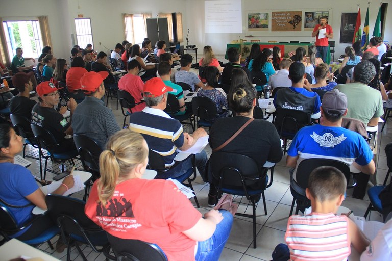 Plateia aparece de costas, enquanto palestrante fala na frente da sala