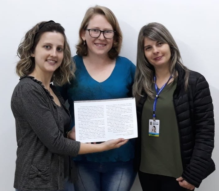 Foto de três mulheres olhando para a câmera, segurando um documento