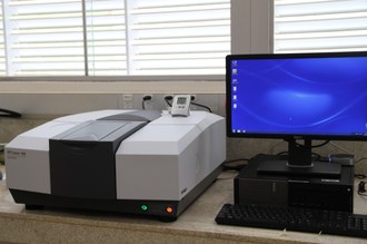 Na imagem, o espectrofotômetro está posicionado em uma bancada, ao lado de um computador.