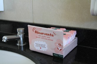 Foto de uma bancada de banheiro. Sobre a bancada uma caixa em tons de rosa contém absorventes dentro dela. Na frente da caixa aparecem os dizeres: Absorvente para ajudar você, quando você mais precisa! Lembre-se que, assim como você, outra colega pode precisar. use com consciência.