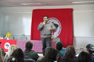 Na imagem uma pessoa em pé, com microfone na mão, fala para a plateia que está sentada em auditório.