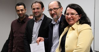Na imagem quatro pessoas estão em pé, posando para foto. Uma delas que está ao centro segura em suas mãos documento assinado.