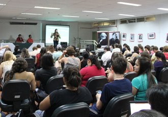 Na imagem pessoas sentadas em auditório acompanham apresentação de palestrante que está no palco.