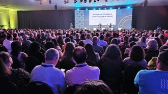 A imagem mostra um auditório lotado, com muitas pessoas sentadas assistindo a uma apresentação em um palco ao fundo. No palco, há três pessoas participando do evento: uma está em pé apresentando algo e outras duas estão sentadas, participando da conversa.  O telão ao fundo exibe o título da palestra: "Emergência Climática e Caminhos para os futuros!", seguido do nome da palestrante, Paula Netto, PhD, e a instituição que ela representa, o Instituto Nacional de Pesquisas Espaciais - INPE. Também aparecem os logotipos de Itaipu Binacional e MCTI (Ministério da Ciência, Tecnologia e Inovação), indicando os organizadores ou apoiadores do evento.