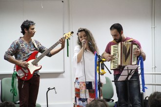 Na imagem três pessoas estão no palco realizando apresentação musical.