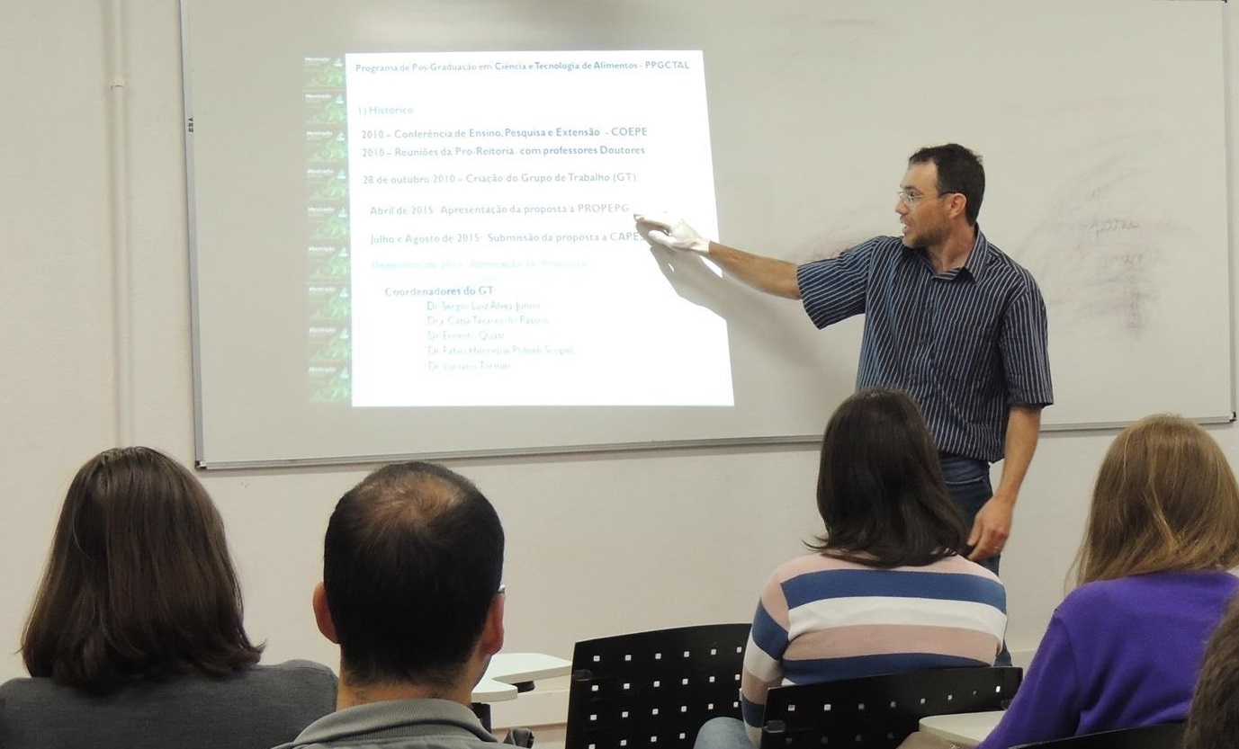 Professor aponta para conteúdo projetado em tela em frente aos alunos.