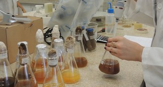 Na imagem são observados várias vidrarias de laboratório sobre uma bancada e a mão de uma mulher manuseando equipamentos.