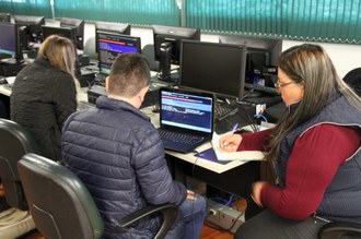 Na imagem dois estudantes sentados utilizam computadores, ao lado deles uma professora passa orientações e faz anotações em caderno.