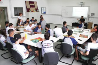 Alunos em sala de aula e professor, ao fundo, explicando o conteúdo.