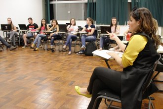Da esquerda para a direita, alunos em suas carteiras, em semicírculo; ao final professora explicando conteúdo.