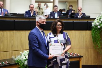 Um homem entrega uma placa para uma mulher. Os dois sorriem para a foto.