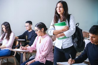 imagem de uma menina de pé carregando livros. Ao seu redor outras pessoas estão sentadas em mesas escolares escrevendo