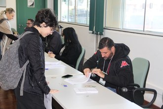 Foto de uma menina de pé em frente a uma mesa. Atrás da mesa um homem sentado manuseia algumas folhas.