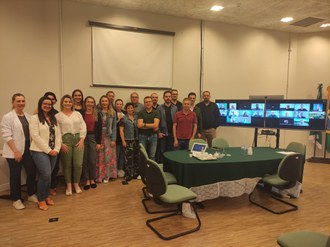 Homens e mulheres de pé posando para a foto. Ao lado televisores e neles imagens de pessoas conectadas em videoconferência