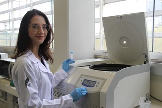 mulher de jaleco branco segura um tubo de ensaio na frente de uma centrífuga de laboratório