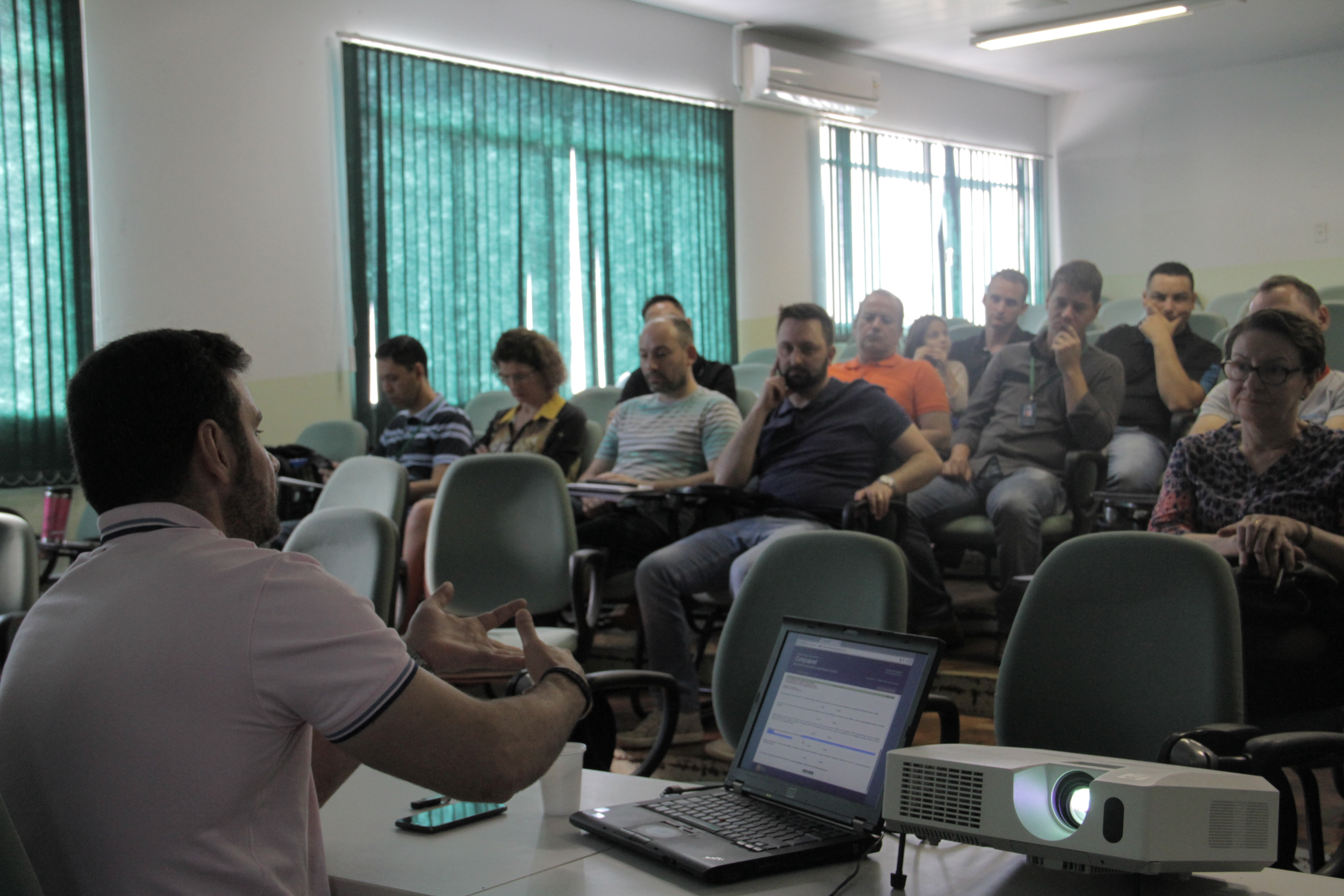 Alt: O palestrante gesticula da mesa central para os participantes do curso. A sua direita, um computador e um projetor multimídia.